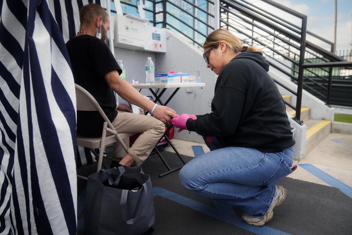A healthcare worker in pink gloves kneels beside a seated patient outside a clinic, treating the person's hand.