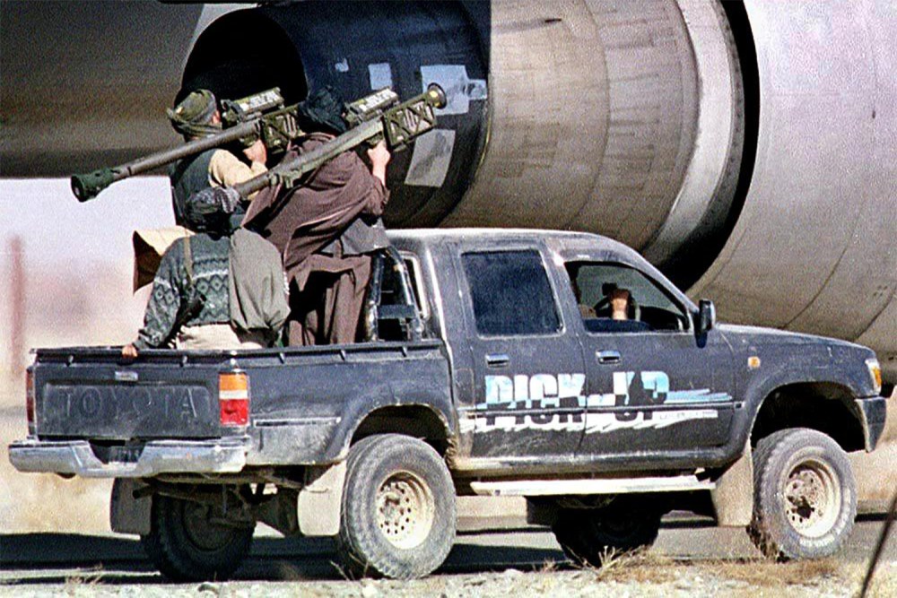 Men standing in the bed of a black pickup truck, aiming shoulder-fired rocket launchers near the jet engine of a large parked aircraft.