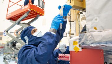 People in blue protective suits and a red cherry picker work in an industrial high bay.