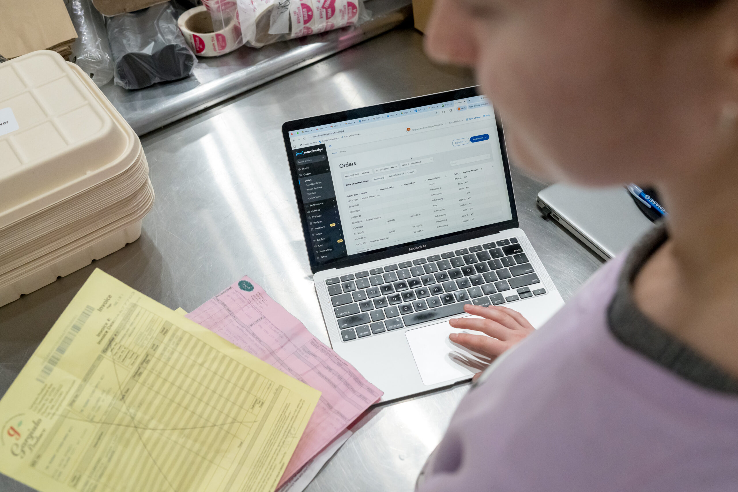 Person using a laptop displaying an order management dashboard, with paperwork and containers on a stainless steel table.