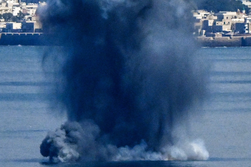 A huge plume of dark smoke coming from a bomb in the the ocean. A city can be seen in the background. 
