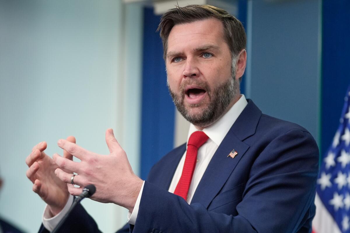 A man with dark hair and beard, in a dark blue suit and red tie, gestures with his hands while speaking 