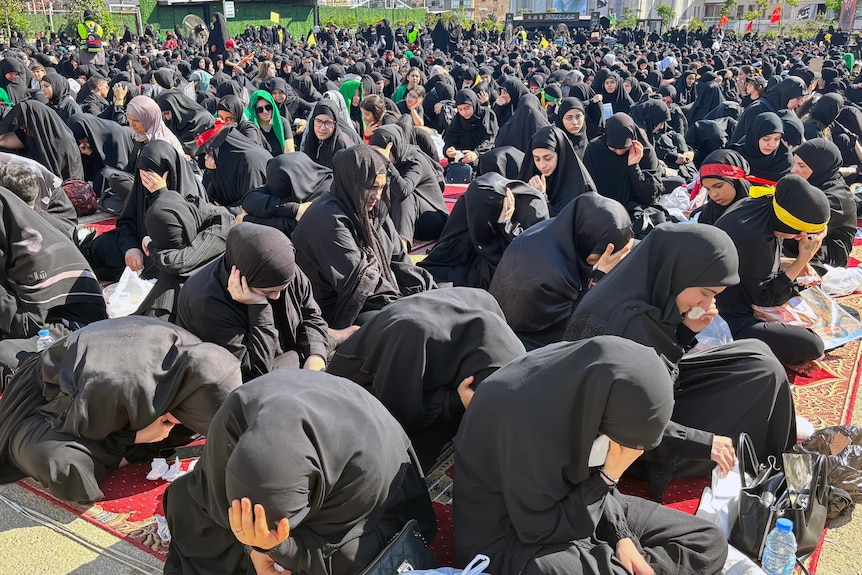 Dozens of women wearing black hijabs and robes are seen gathered and sitting on the ground, crying.