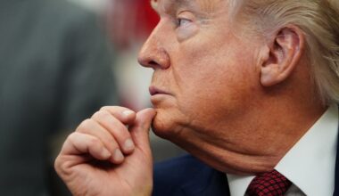 Donald Trump with hand on chin, appearing contemplative during a public event, wearing a suit and red tie.
