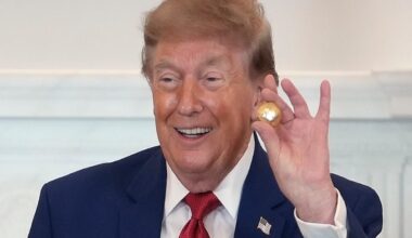 Donald Trump smiling and holding a small golden ball, wearing a blue suit and red tie.