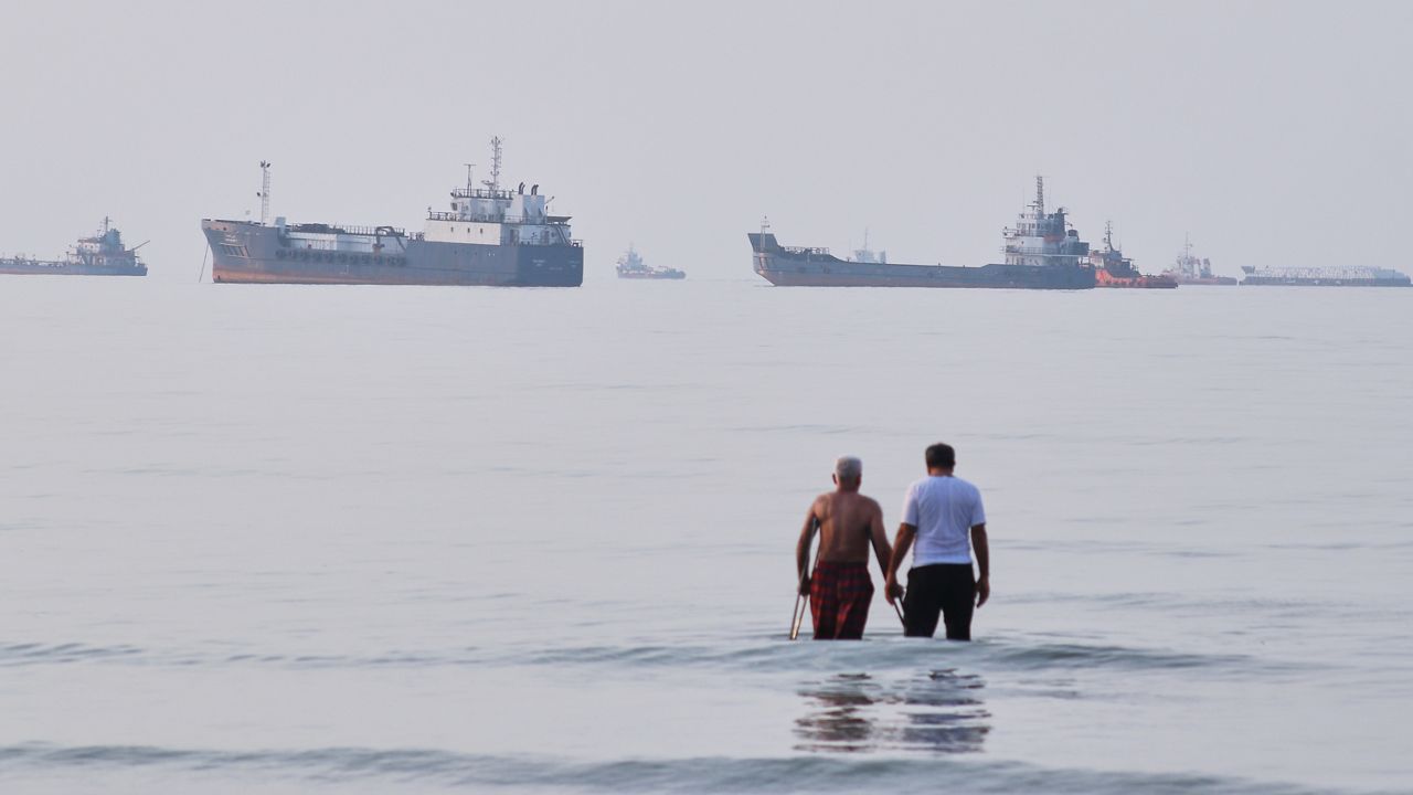 Two men wade in the waters of the Strait of Hormuz with vessels anchored in the background, off Bandar Abbas, Iran, Sunday, July 12, 2026. (Razieh Poudat/ISNA via AP)