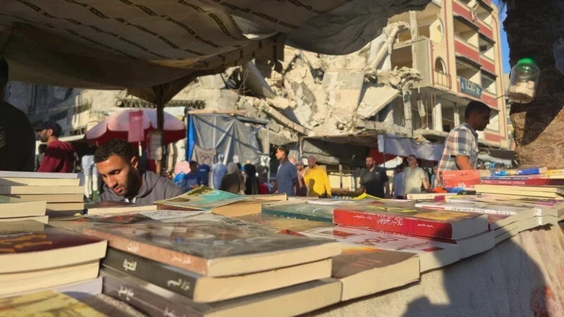 beneath a tent a collection of books is displayed for sale in a bustling market with a partially collapsed building in the background