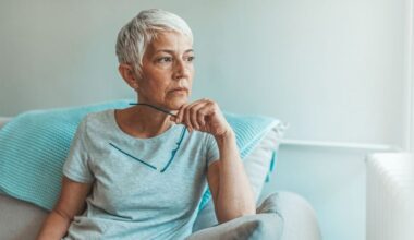 Donna di mezza età con i capelli bianchi e corti seduta sul divano con gli occhiali in mano che guarda fuori dalla finestra
