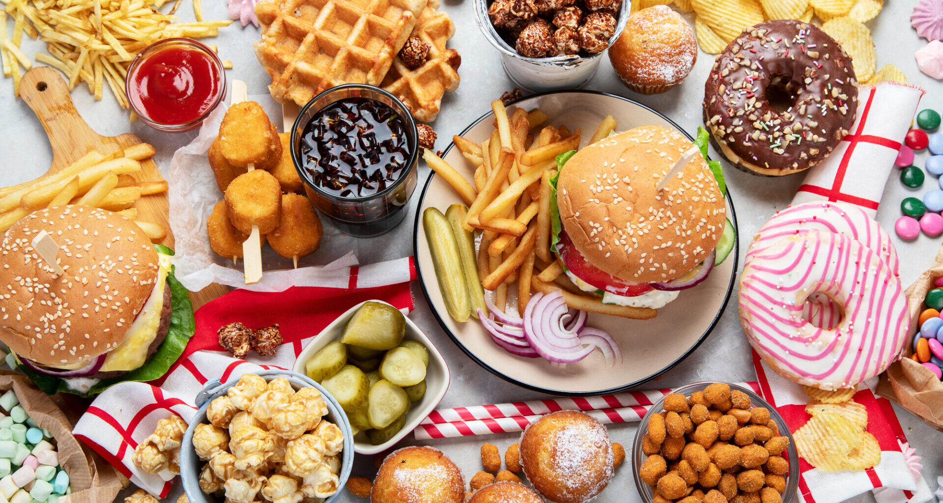 Assortimento di cibo spazzatura visto dall'alto: panini con hamburger, nuggets, pop corn, patatine, waffle, donut glassati, cupcake, marshmallow, ketchup; concept: snack, junk food, ultra processati