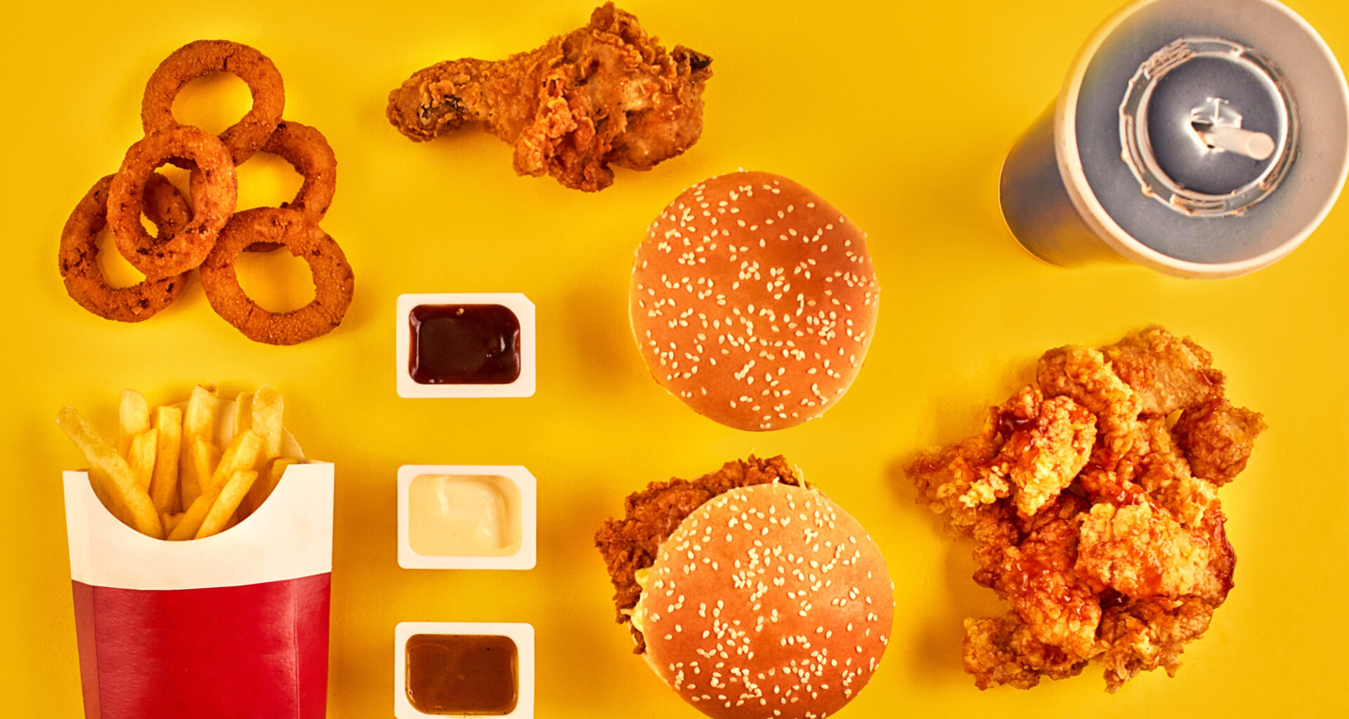 Alcuni alimenti da fast food visti dall'alto su una superficie gialla: panini con hamburger, pollo fritto, anelli di cipolla, patatine , bibita zuccherata e salse; concept: junk food, ultra processati