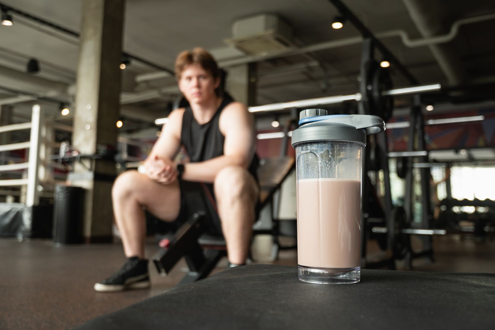 Protein shake in foreground and blurred athlete seated in gym focusing on recovery. Workout at modern gym.