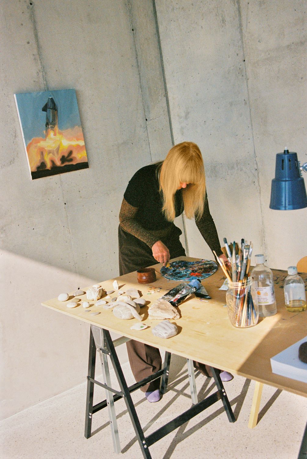 Goshka Macuga al tavolo da lavoro, tavolo in legno con diversi pennelli, lampada blu e quadro appeso alla parete