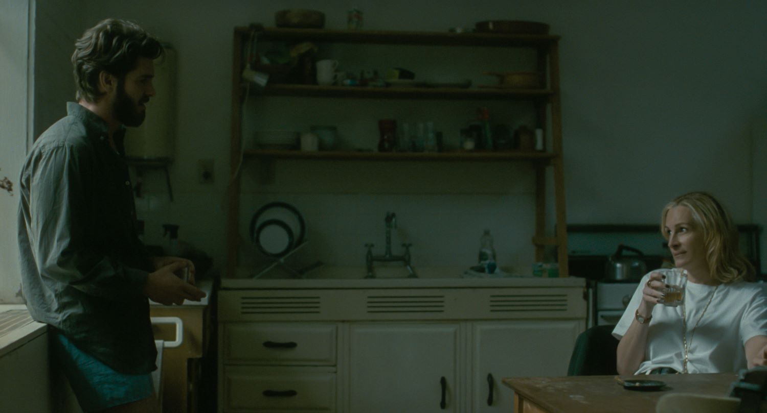 Uomo e donna in cucina, lei seduta al tavolo con un bicchiere in mano, lui in piedi rivolto verso di lei, la cucina è bianca e beige