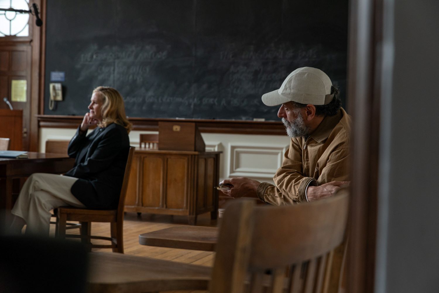 Uomo e donna seduti in un aula scolastica su sedie di legno, sullo sfondo una cattedra di legno e una grossa lavagna nera sporca di gesso appesa alla parete