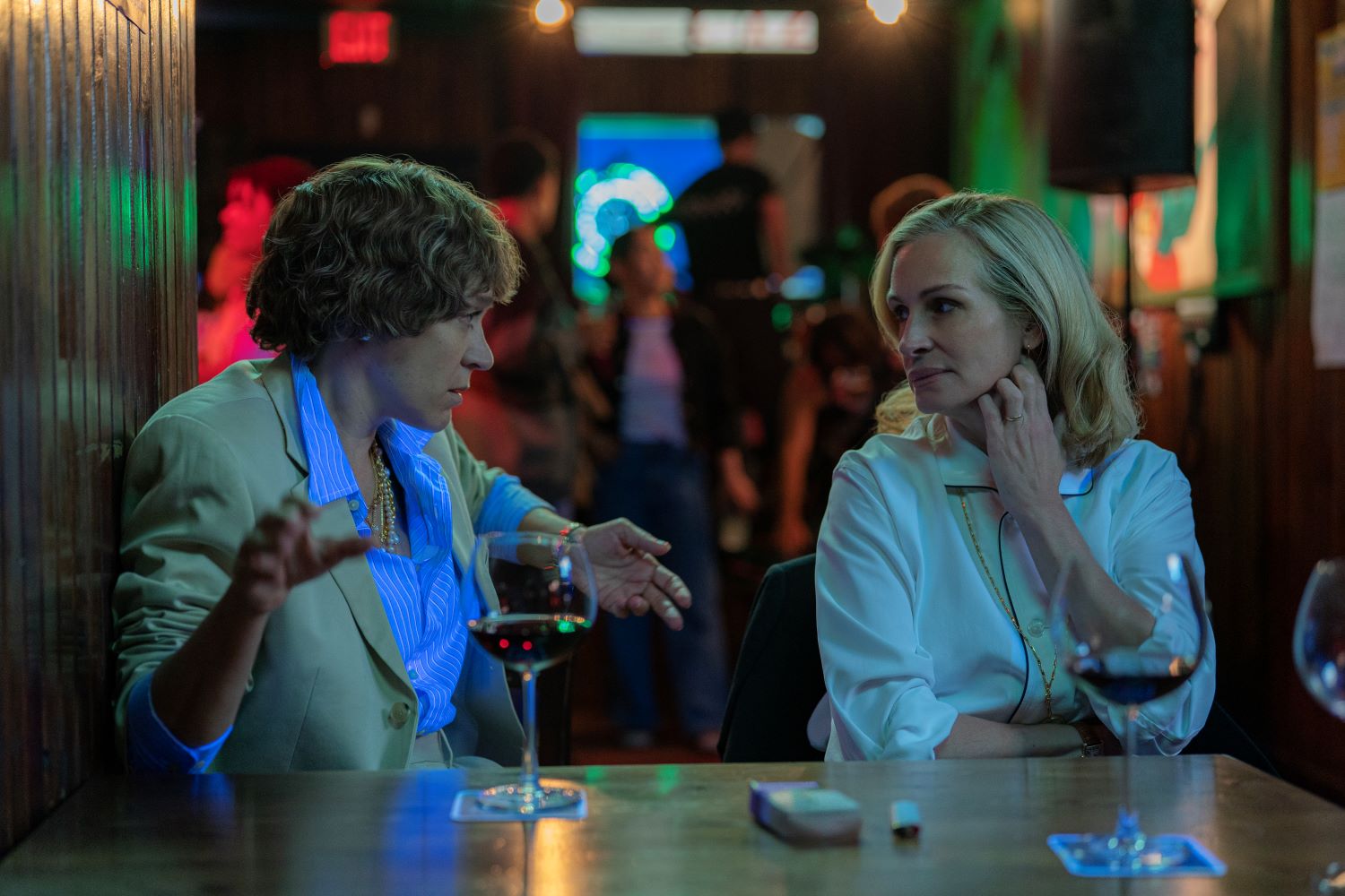 Due donne sedute ad un bar che parlano, un tavolo di legno sul quale sono appoggiati due bicchieri di vino, sullo sfondo luci e persone all'interno del locale