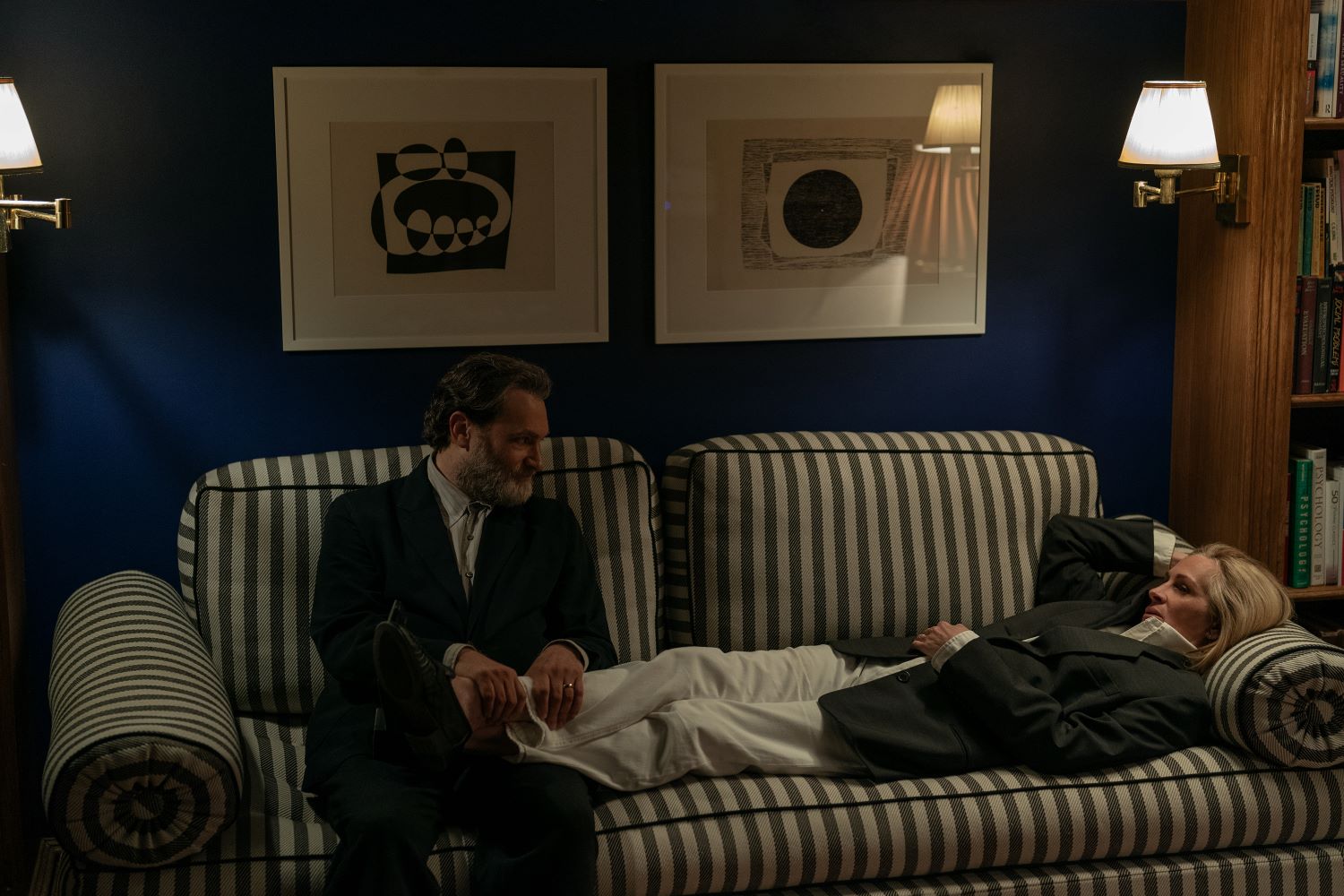 Un uomo e una donna su un divano a righe bianco e grigio, pareti blu e due grossi quadri appesi sopra il divano