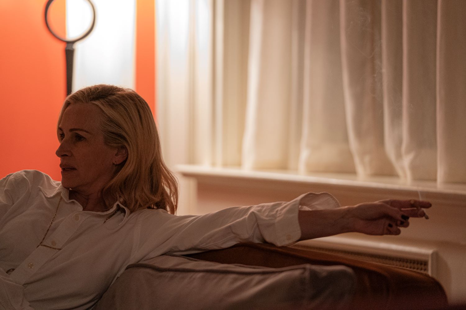 Donna bionda seduta sul divano, sullo sfondo tende bianche e una parete arancione