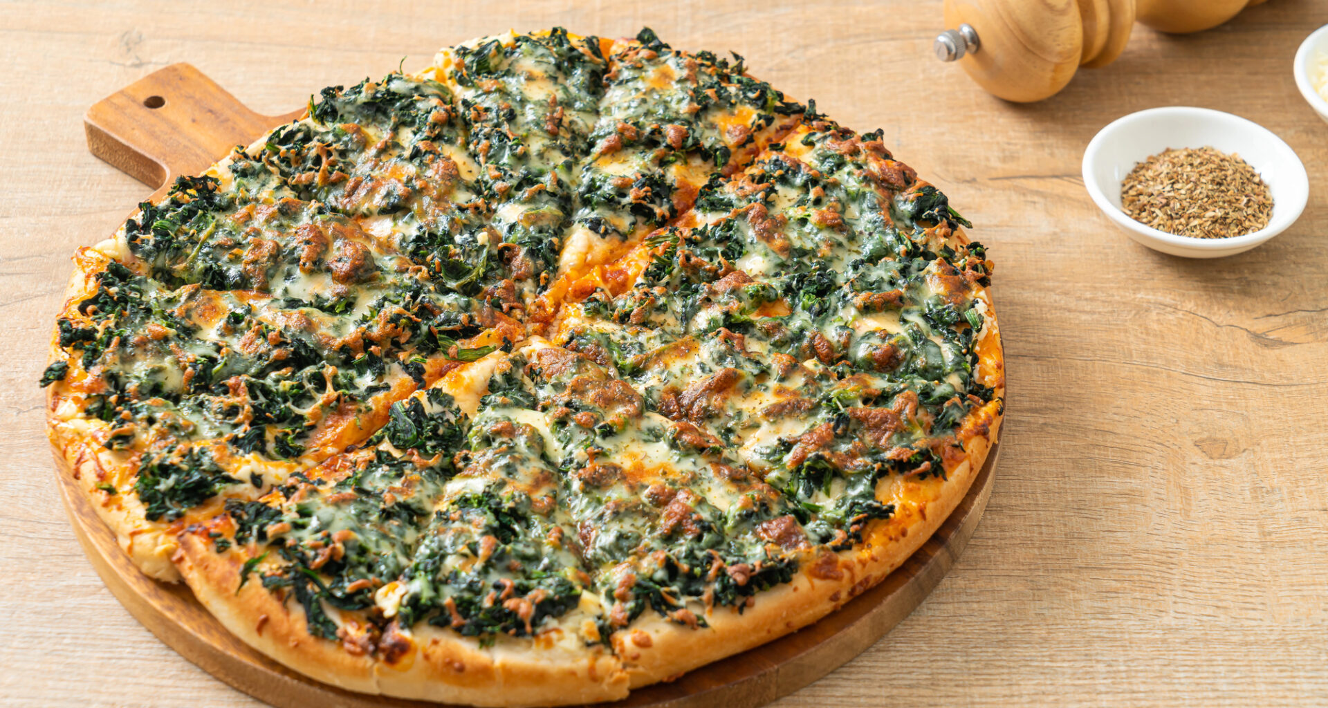 Una pizza con spinaci o friarielli, salsiccia e formaggio su un tagliere rotondo, tagliata a fette, sullo sfondo macina pepe e ciotola di pepe