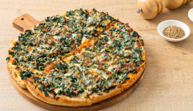 Una pizza con spinaci o friarielli, salsiccia e formaggio su un tagliere rotondo, tagliata a fette, sullo sfondo macina pepe e ciotola di pepe