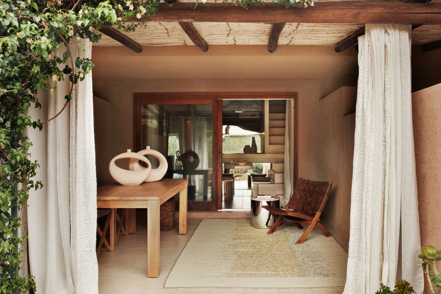 piccolo patio con pergola in cannicciato, pareti marroncine, tappeto in lana, appeso al muro una cesta di giunco e rafia, tavolo di legno con due vasi bianchi e tende bianche