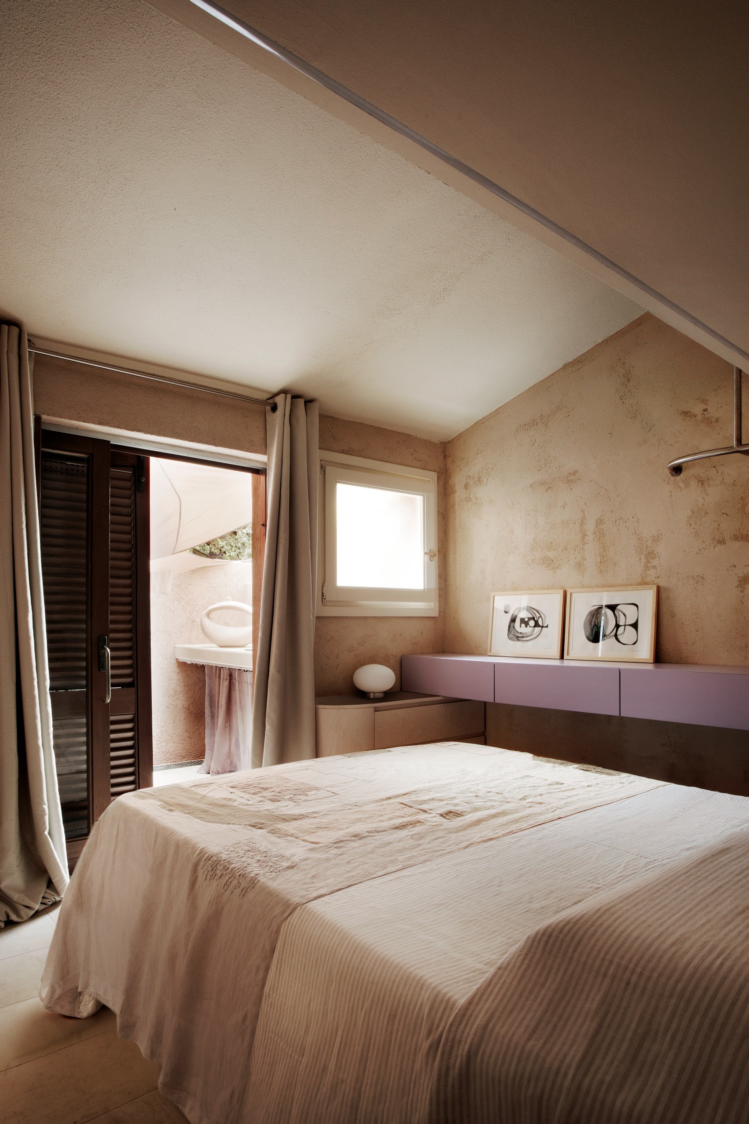 camera da letto con pareti bige, lenzuola bianche, mobili lilla e beige, quadri e vasi bianchi, piccolo balcone