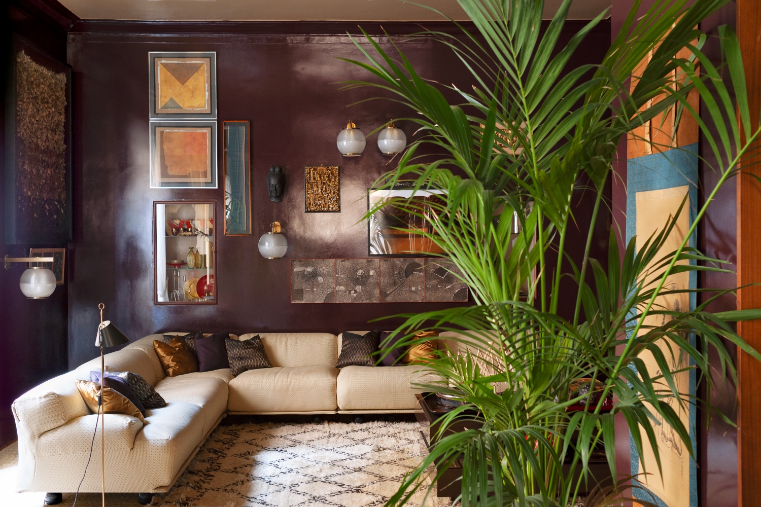 Zona living con divano modulare beige, pareti marroni con quadri appesi, pianta verde alta, applique bianche e tappeto beige