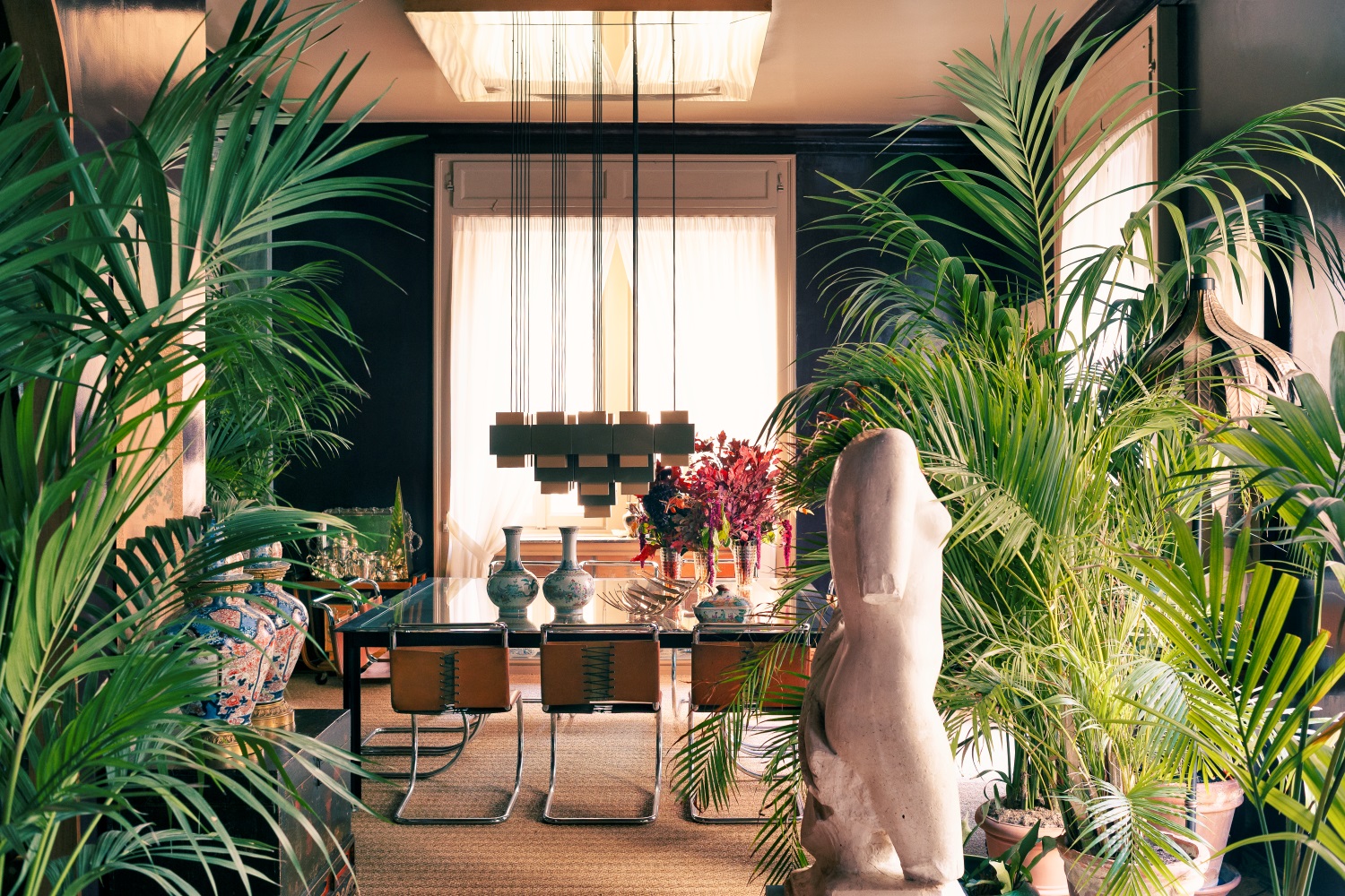 Sala da pranzo con tavolo in vetro, una statua di un busto, diverse piante verdi, vasi colorati, sedie marroni in pelle