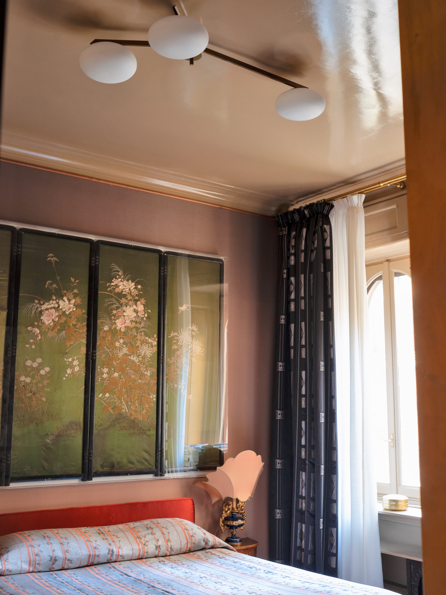 Camera da letto con pareti marroncine, una finestra con tende bianche e blu, copriletto azzurro e rosso, lampada da tavolo sul comodino, lampada da soffitto bianca