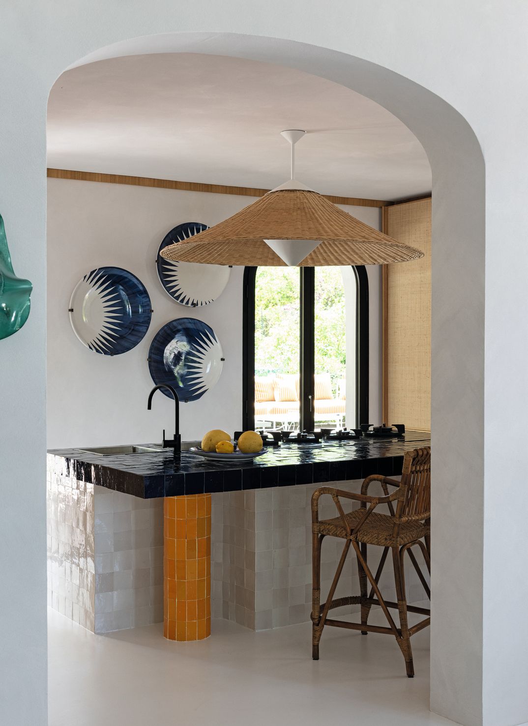 Cucina isola rivestita di piastrelle blu bianche gialle lampadario in vimini sgabello in vimini piatti alle pareti finestra