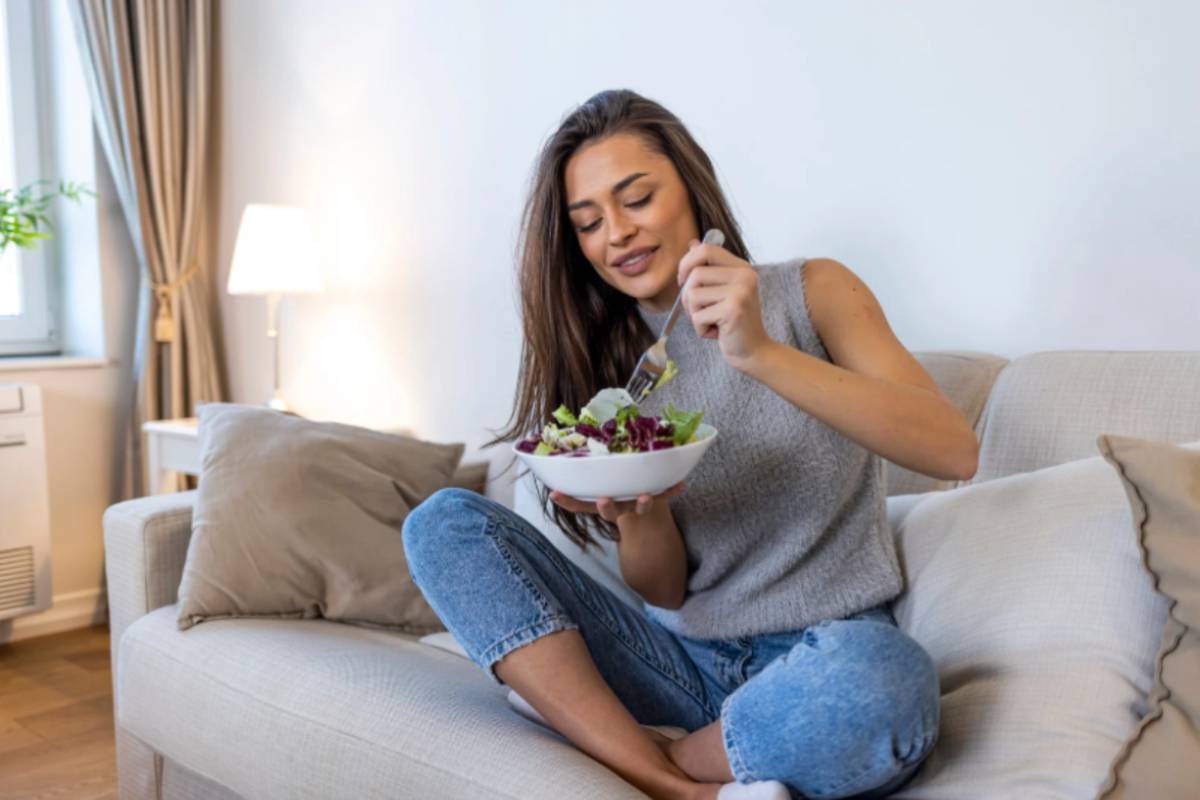 donna seduta sul divano che mangia un piatto di insalata