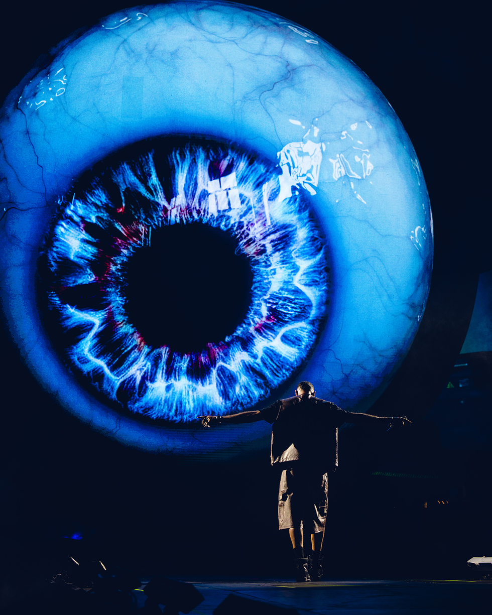 a performer stands with arms outstretched in front of a large glowing blue eye display