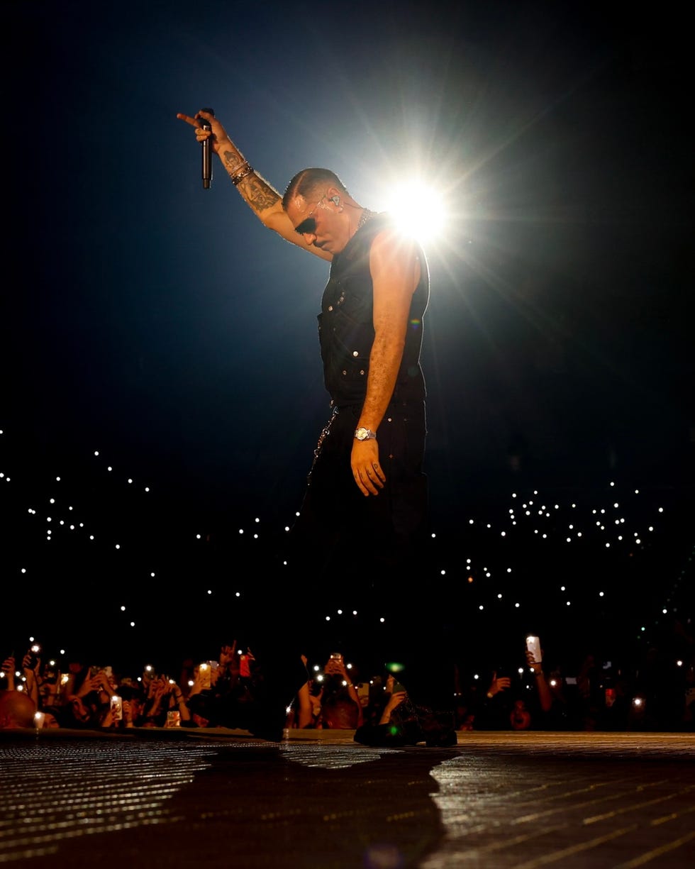a performer on stage raising a microphone with a bright light behind them and an audience illuminated by cellphone lights