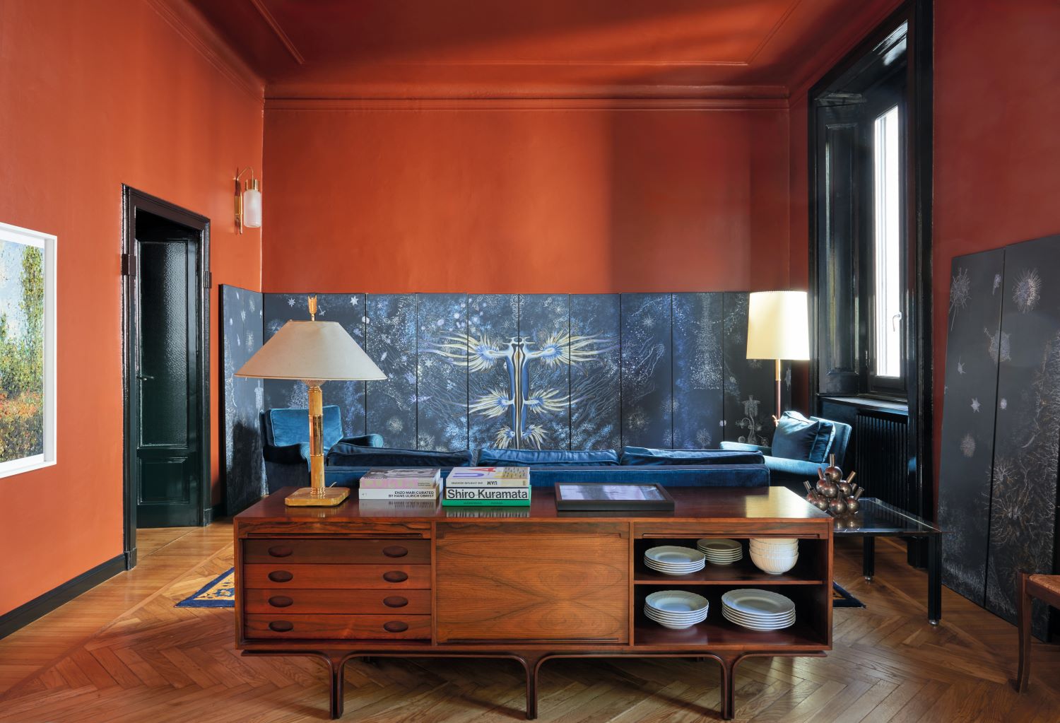 Soggiorno con madia in legno, parquet, divani blu, soffitto e pareti rosse, applique e due lampade da terra e da tavolo