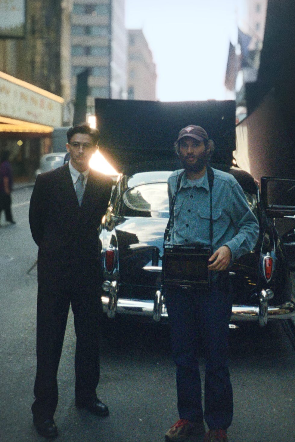 A man in a suit and a man holding a camera stand in front of a vintage car.