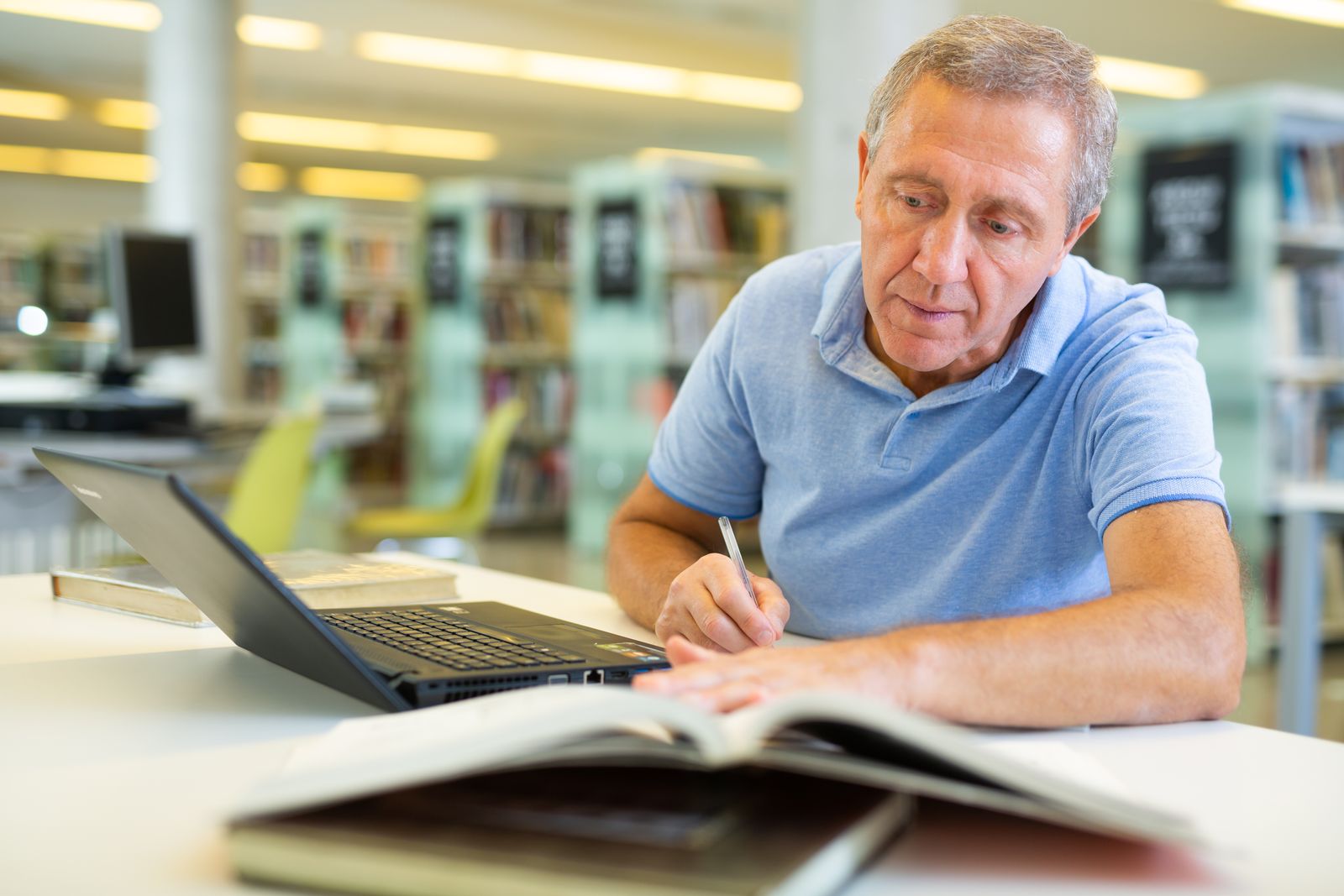 Concentrated mature man spending time in library reading books and making laptop. Selfeducation concept