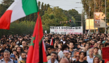A photograph of a street in the Pilastro neighbourhood of Bologna on 26 July 2026, thronged with people marching in protest about the death of Abderrahim Fakir. Some are holding a large white banner with the