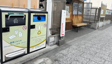 Two bins for waste disposal beside a wooden bench at a bus stop.