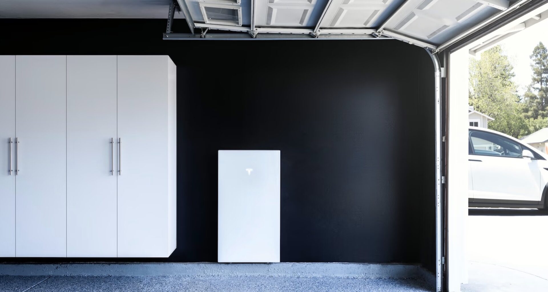 A garage featuring white cabinets, a Tesla Powerwall, and a partially open door revealing a car outside.