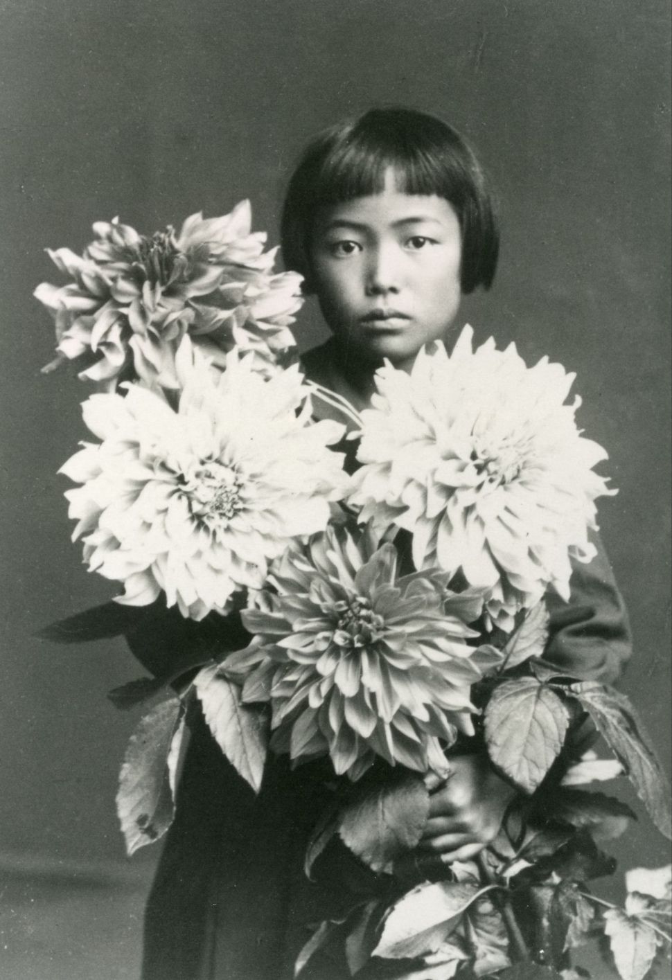 A black-and-white childhood portrait of Yayoi Kusama posing behind a large arrangement of flowers.