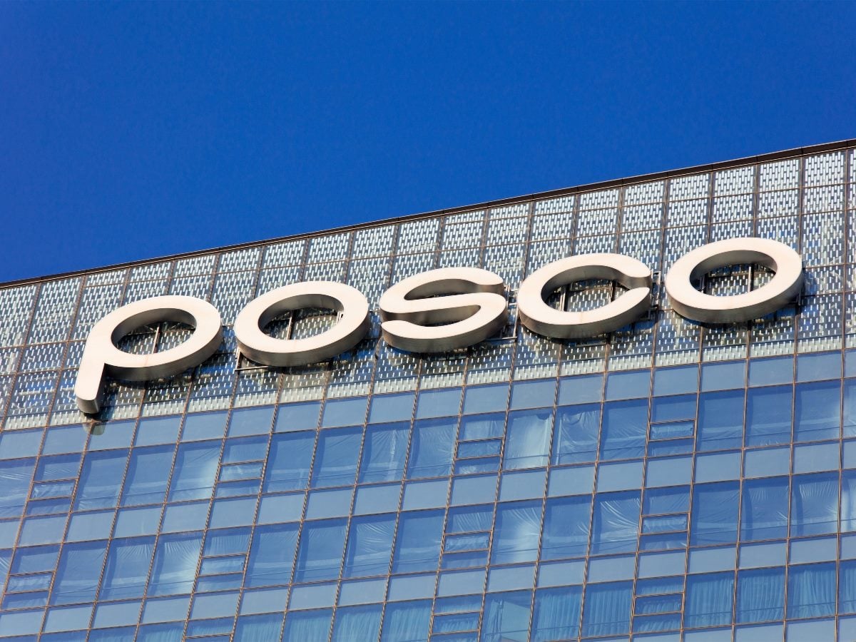 Close-up view of the POSCO logo displayed in large silver three-dimensional letters mounted on the upper facade of a modern glass office tower, shot from a low angle against a clear deep blue sky, with the building's grid pattern of windows and perforated metal paneling visible beneath the sign.