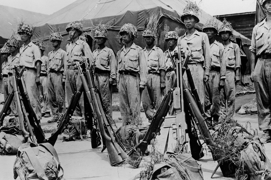 A black and white photo of men standing to attention in rows behind guns placed together in a pyramid.