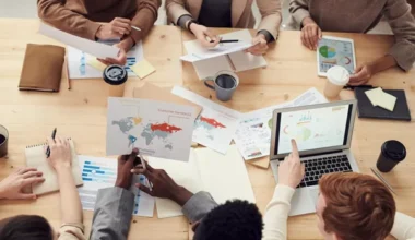 Grupė žmonių sėdi prie didelio medinio stalo, ant kurio išdėlioti dokumentai su grafikais, nešiojamasis kompiuteris ir kavos puodeliai; jie diskutuoja ir planuoja strategiją.