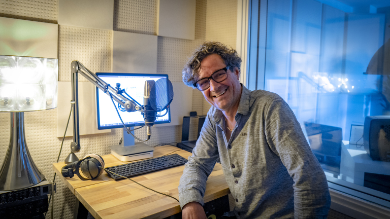 Man met krullend haar en bril zit glimlachend voor een microfoon in een geluidsstudio naast een computer en hoofdtelefoon.