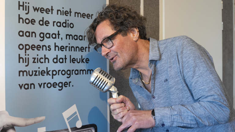 Een man met krullend haar en een bril spreekt in een retro microfoon naast een blauwe poster met Nederlandse tekst over het herinneren van een muziekprogramma van vroeger.