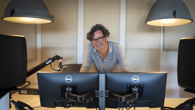 Man met krullend haar en bril staat achter twee Dell-monitoren in een verlicht kantoor met een microfoon op een statief.