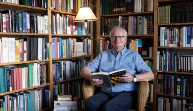 Foto van een oudere man met grijs haar en bril. Hij zit op een rieten stoel in een hoek met achter hem kasten vol boeken. Hij draagt een lichtblauw overhemd en een donkerblauwe broek. HIj houdt een opengeslagen boek in zijn handen.