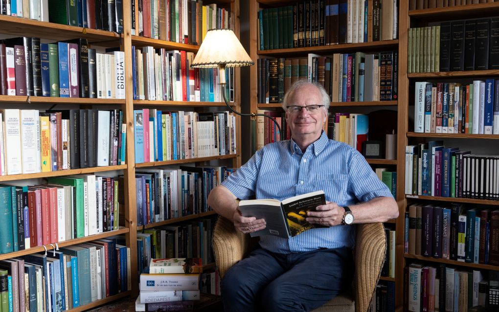 Foto van een oudere man met grijs haar en bril. Hij zit op een rieten stoel in een hoek met achter hem kasten vol boeken. Hij draagt een lichtblauw overhemd en een donkerblauwe broek. HIj houdt een opengeslagen boek in zijn handen.