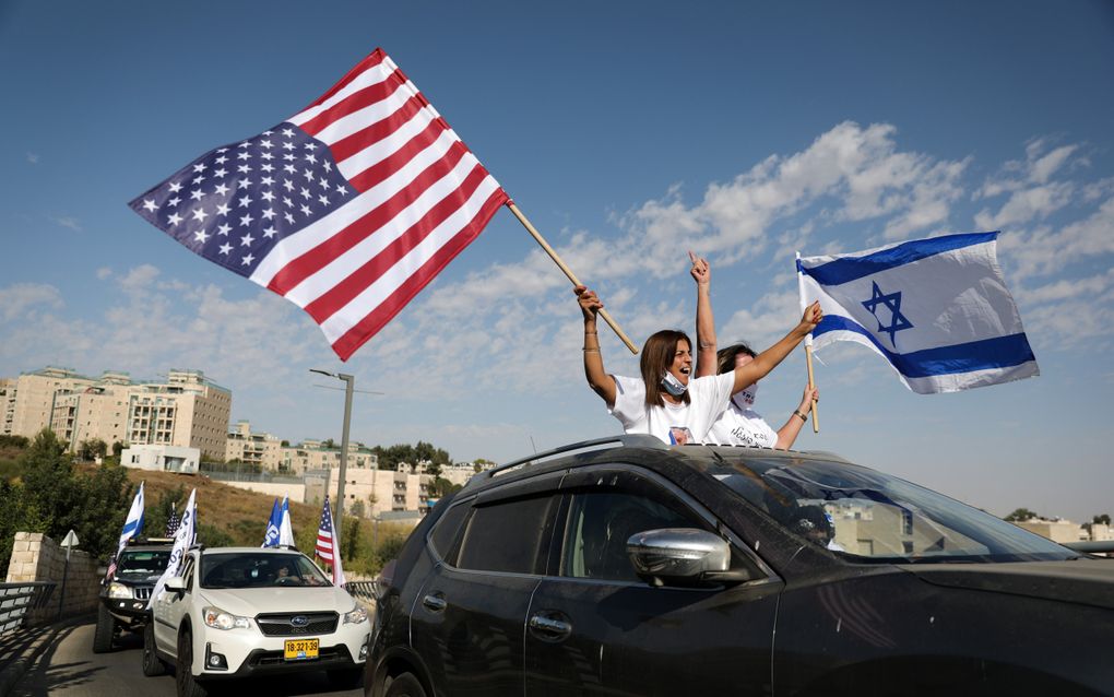 Twee vrouwen in wit T-shirt zwaaien met vlaggen vanuit een auto.