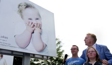 Drie mensen, waarvan twee met Downsyndroom, staan bij een groot billboard met daarop een blond lachend jongetje met het syndroom van Down, dat zijn handen voor zijn mond slaat.