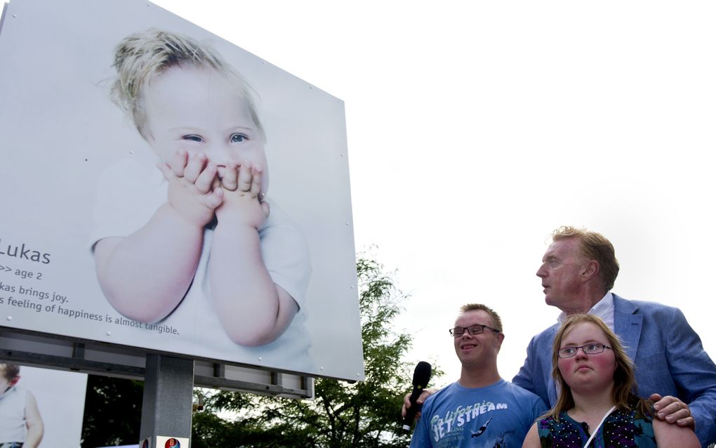 Drie mensen, waarvan twee met Downsyndroom, staan bij een groot billboard met daarop een blond lachend jongetje met het syndroom van Down, dat zijn handen voor zijn mond slaat.